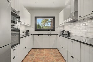 A modern kitchen with white cabinetry, a black countertop, a window with a view of trees, and a tiled backsplash. - Villa 'Can Ayala' Arenys de Munt – With Garden and Private Pool. (Collsacreu)