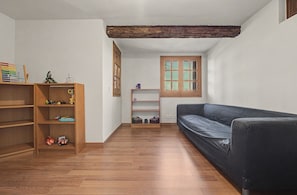 A room with a wooden floor, a black sofa, a wooden bookshelf, and a window with a wooden frame. - Villa 'Can Ayala' Arenys de Munt – With Garden and Private Pool. (Collsacreu)