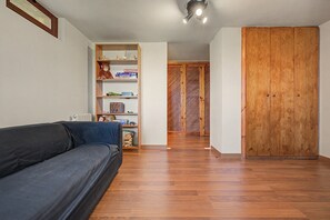 Una sala de estar con un sofá oscuro, piso de madera, una estantería con diversos objetos y un armario de madera.