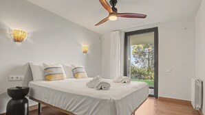 A modern bedroom with a bed, bedside lamps, a ceiling fan, and a view of greenery through a glass door. - Villa 'Can Ayala' Arenys de Munt – With Garden and Private Pool. (Collsacreu)