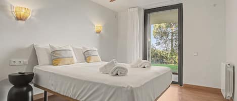 A modern bedroom with a bed, bedside lamps, a ceiling fan, and a view of greenery through a glass door.