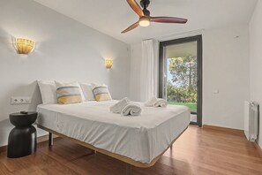Ein modernes Schlafzimmer mit Bett, Nachttischlampen, Deckenventilator und Blick auf Grün hinter einer Glastür.