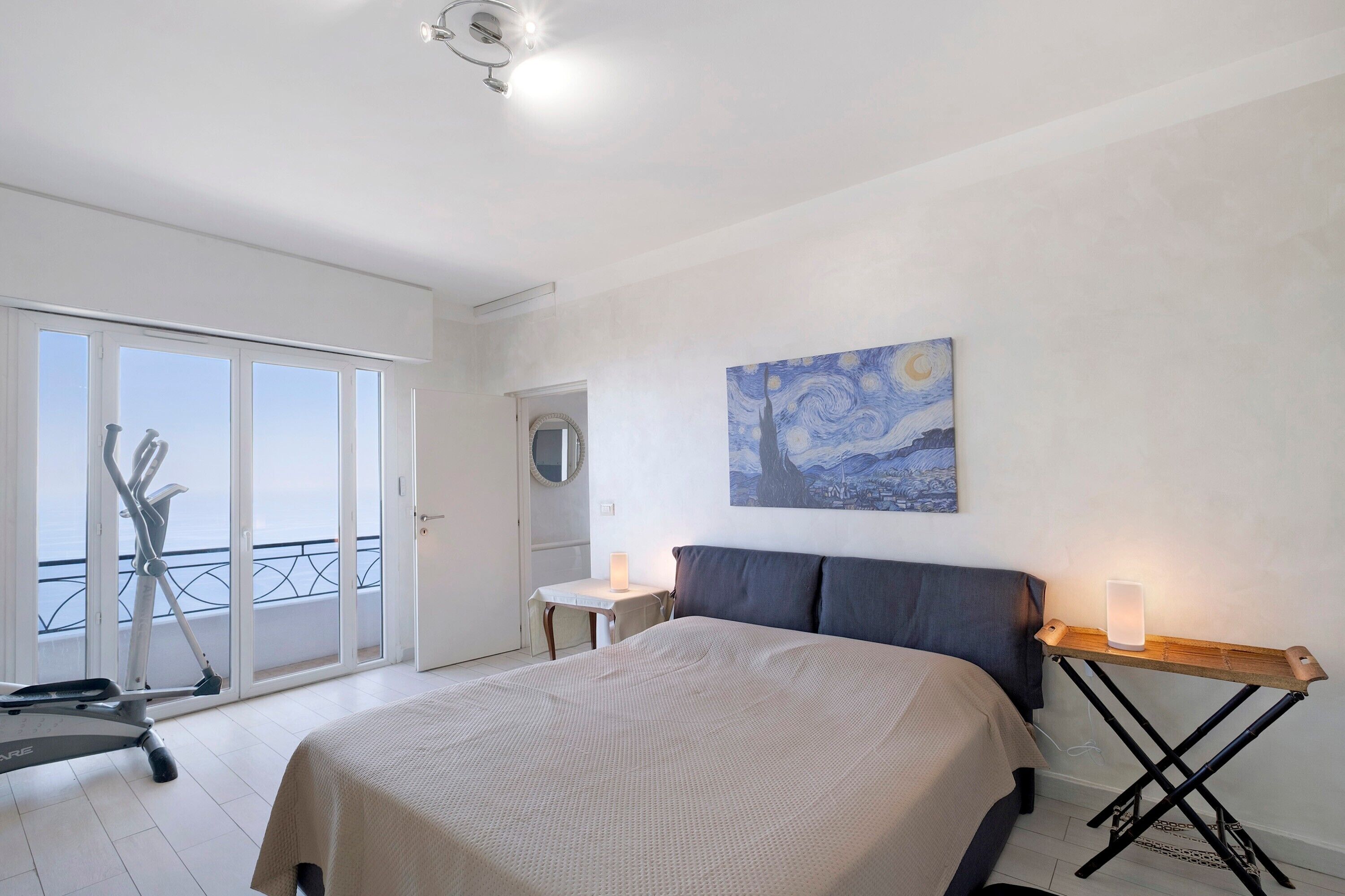 A bedroom with a bed, a desk, a standing lamp, a painting, and a balcony with exercise equipment.