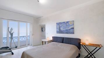 A bedroom with a bed, a desk, a standing lamp, a painting, and a balcony with exercise equipment.