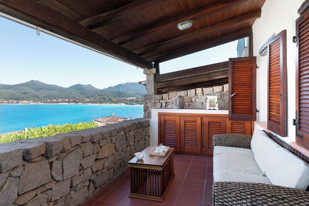 Un balcón con muro de piedra, mobiliario de madera y vista al mar y a las montañas.