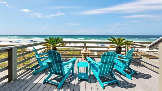 A wooden deck with turquoise Adirondack chairs and a small table, overlooking a beach with umbrellas and people. - Beach Front Townhome in Miramar Beach (Miramar Beach)