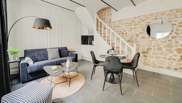 Uma sala de estar moderna com um sofá, mesa de jantar, cadeiras, luminária e uma escada com parede de pedra.