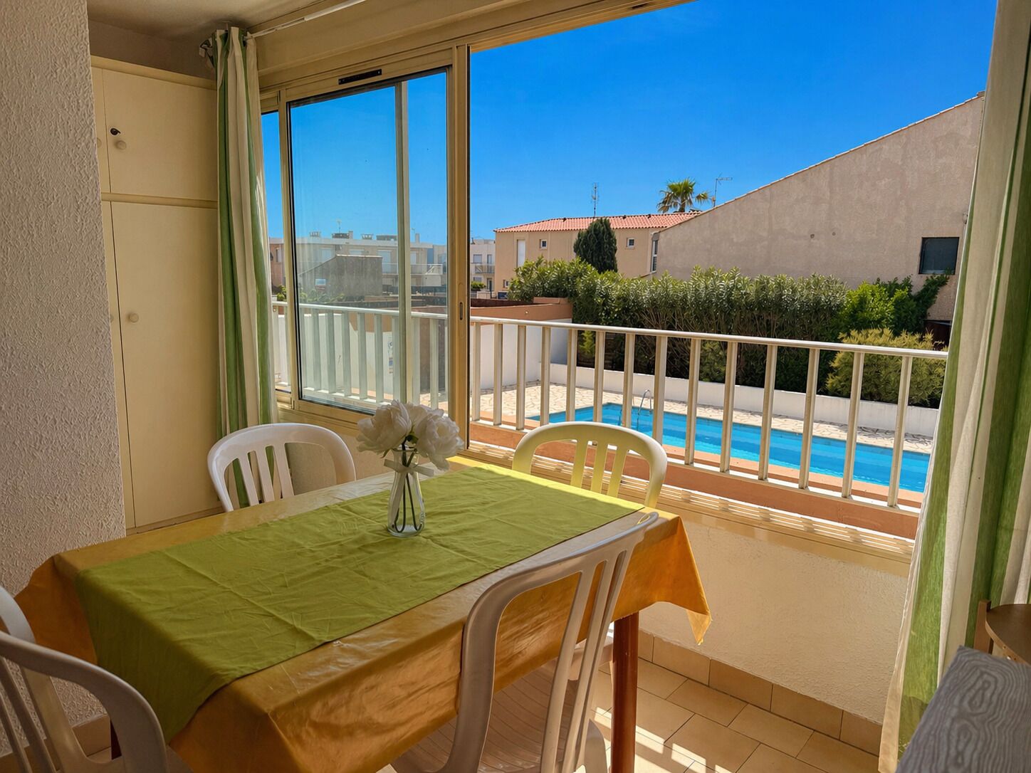Un balcon avec une piscine, des chaises blanches, une table recouverte d’une nappe verte, et une vue sur des bâtiments et la verdure.
