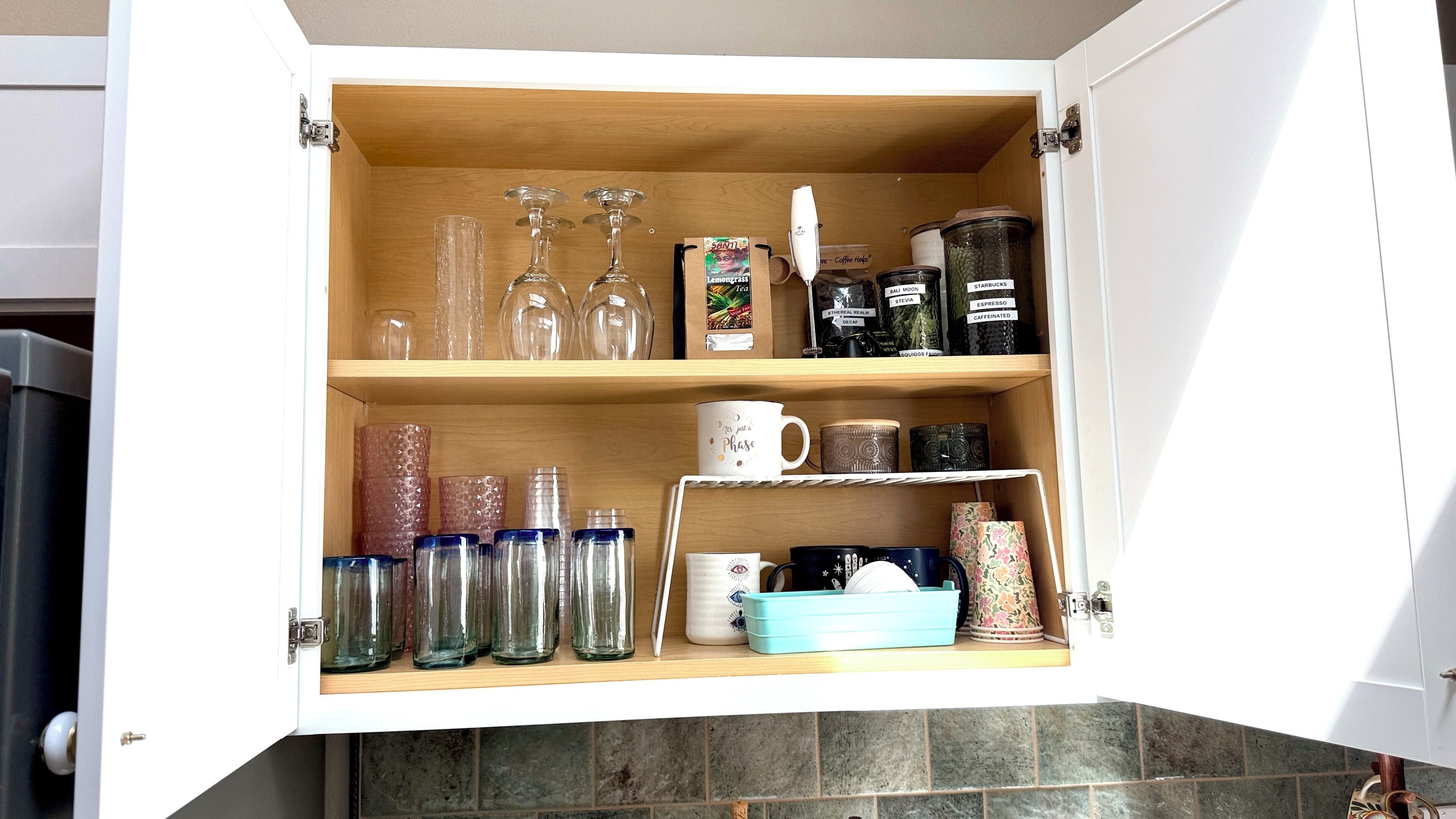 A kitchen cabinet with shelves containing glassware, coffee containers, and a blue dish rack.