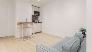 A modern living room with a grey sofa, a wooden dining table with bar stools, and a kitchen area with white cabinets and a microwave. - Txapitela Rooms - Perfect for 2 (Pamplona)