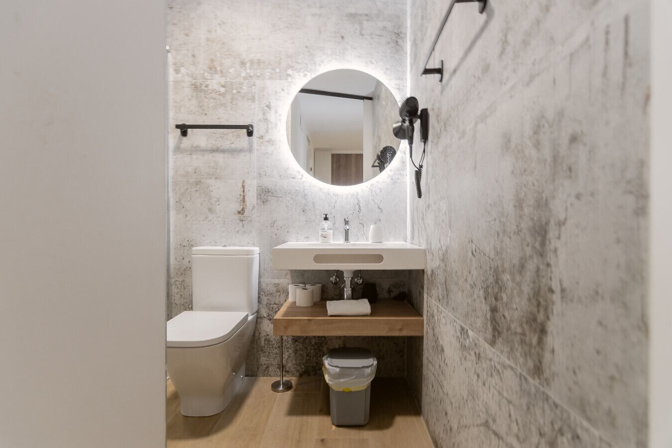 Un baño con inodoro blanco, tocador de madera, espejo con luces y secador de pelo montado en la pared.