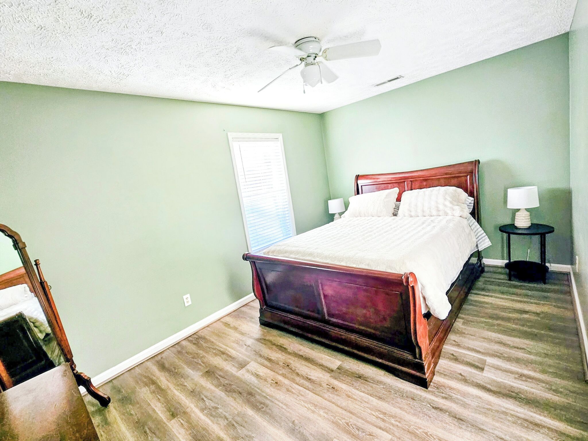 A bedroom with a wooden bed, a ceiling fan, a nightstand with a lamp, and a window with blinds.