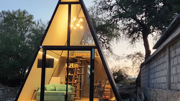 A triangular glass house with a bed, a ladder, and a small table.
