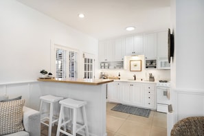 Une cuisine moderne avec des meubles blancs, un plan de travail en bois et une cuisinière intégrée. On y trouve deux tabourets de bar blancs et un petit coin salon avec un canapé gris.