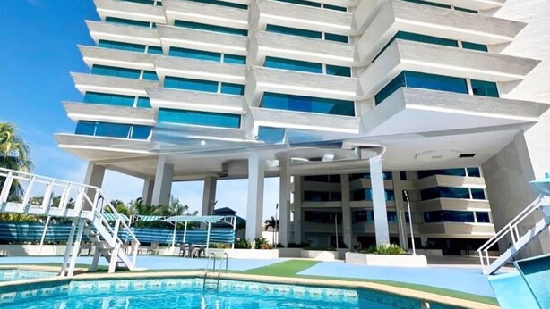 Een modern hotel met een zwembad, meerdere balkons en een helderblauwe lucht.