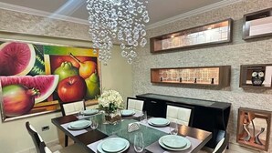Een eetgedeelte met een groot fruit schilderij, een kroonluchter en ingebouwde planken met decoratieve objecten.