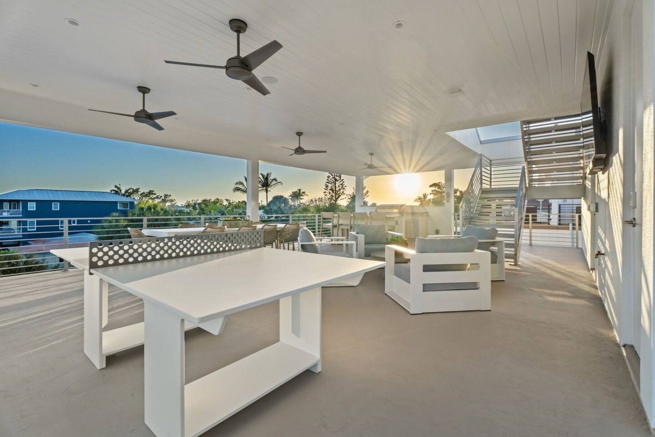 Un espace de vie extérieur moderne, avec une table et des chaises blanches, des ventilateurs de plafond et une vue sur le coucher de soleil.