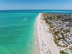 An aerial view of a coastal area with a beach, clear turquoise waters, and a residential area with houses. - Casa Del Sol • Rooftop Deck, Pool & Ocean Views (Anna Maria)