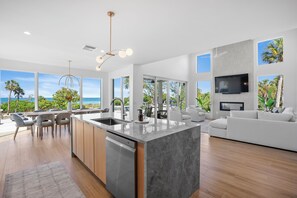 A modern kitchen with a large island, dining area with chairs, a sofa, and a TV mounted on the wall. - Casa Del Sol • Rooftop Deck, Pool & Ocean Views (Anna Maria)