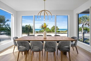 A dining area with a wooden table and grey chairs, large windows with a view of the ocean, and a modern chandelier. - Casa Del Sol • Rooftop Deck, Pool & Ocean Views (Anna Maria)