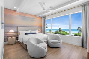 A modern bedroom with a large bed, two armchairs, a ceiling fan, and a view of the ocean. - Casa Del Sol | Rooftop Deck, Pool + Ocean Views (Anna Maria)
