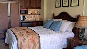 A bedroom with a bed, a nightstand, a lamp, a chair, a ceiling fan, a kitchen area, and framed artwork on the wall.