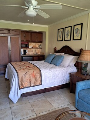 A bedroom with a bed, a nightstand, a lamp, a chair, a ceiling fan, a kitchen area, and framed artwork on the wall. - ISLANDER ON THE BEACH #256
Spectacular sunrises just a few steps to the Ocean!
 (Kapaʻa)