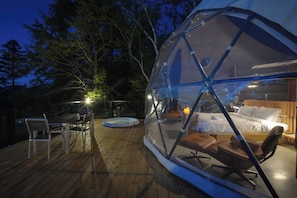Estructura aboveda transparente con una cama, una butaca y una mesa de comedor sobre una terraza de madera.