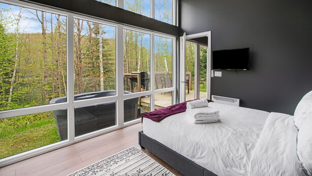 A bedroom with a large window, a bed, a flat-screen TV, and a view of a deck and trees.