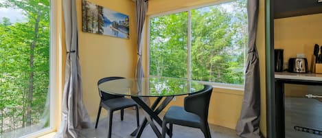 Een kamer met een glazen tafel en stoelen, een schilderij aan de muur en uitzicht op bomen door grote ramen.