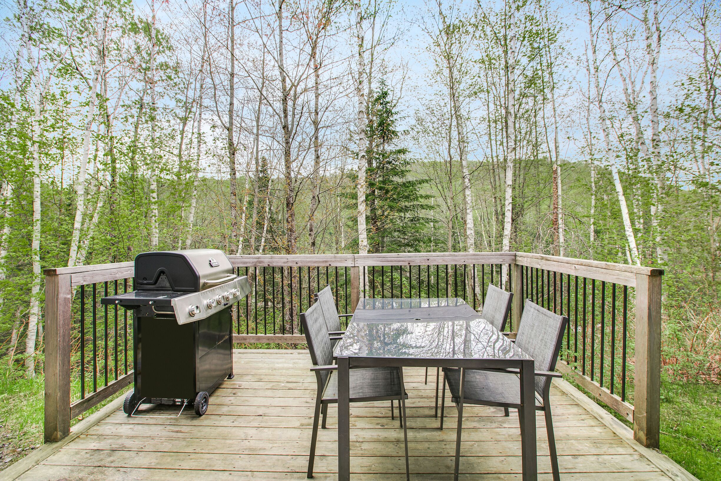 An outdoor deck with a grill, table, and chairs, surrounded by trees.