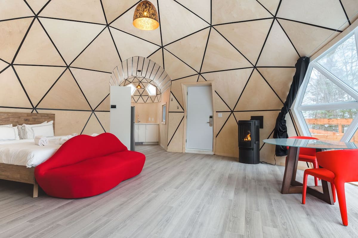 Un dormitorio moderno con diseño geométrico en la pared, una cama, un sillón de peluche rojo, una mesa con una silla y una chimenea.