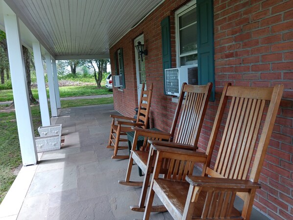 A covered porch with wooden rocking chairs, a brick building, and a car parked outside.