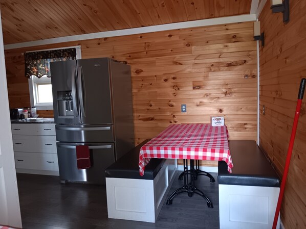 A compact kitchen with a refrigerator, a small table with a checkered tablecloth, wooden walls, and a window with curtains.