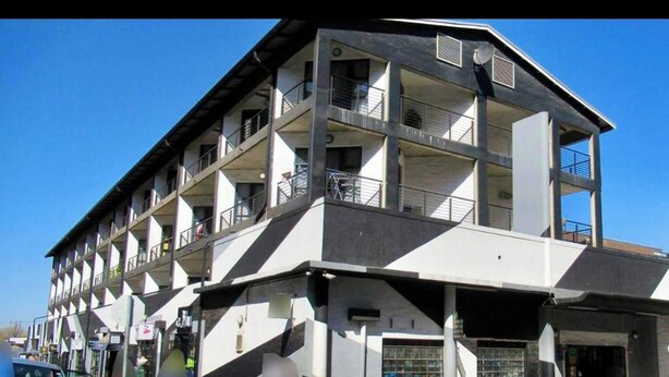 Ein mehrstöckiges Gebäude mit Balkonen, einer flachen Dachterrasse und einem Eingang im Erdgeschoss.