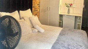 Ein Schlafzimmer mit Ziegelwand, einem Bett mit weißer Bettwäsche, einem schwarzen Ventilator, einem Kleiderschrank und einem kleinen Tisch mit einer Vase.