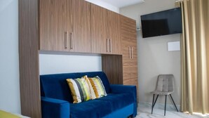 A room with a blue sofa, wooden cabinets, a TV, and a chair. - Augustea Travel (San Marcellino)