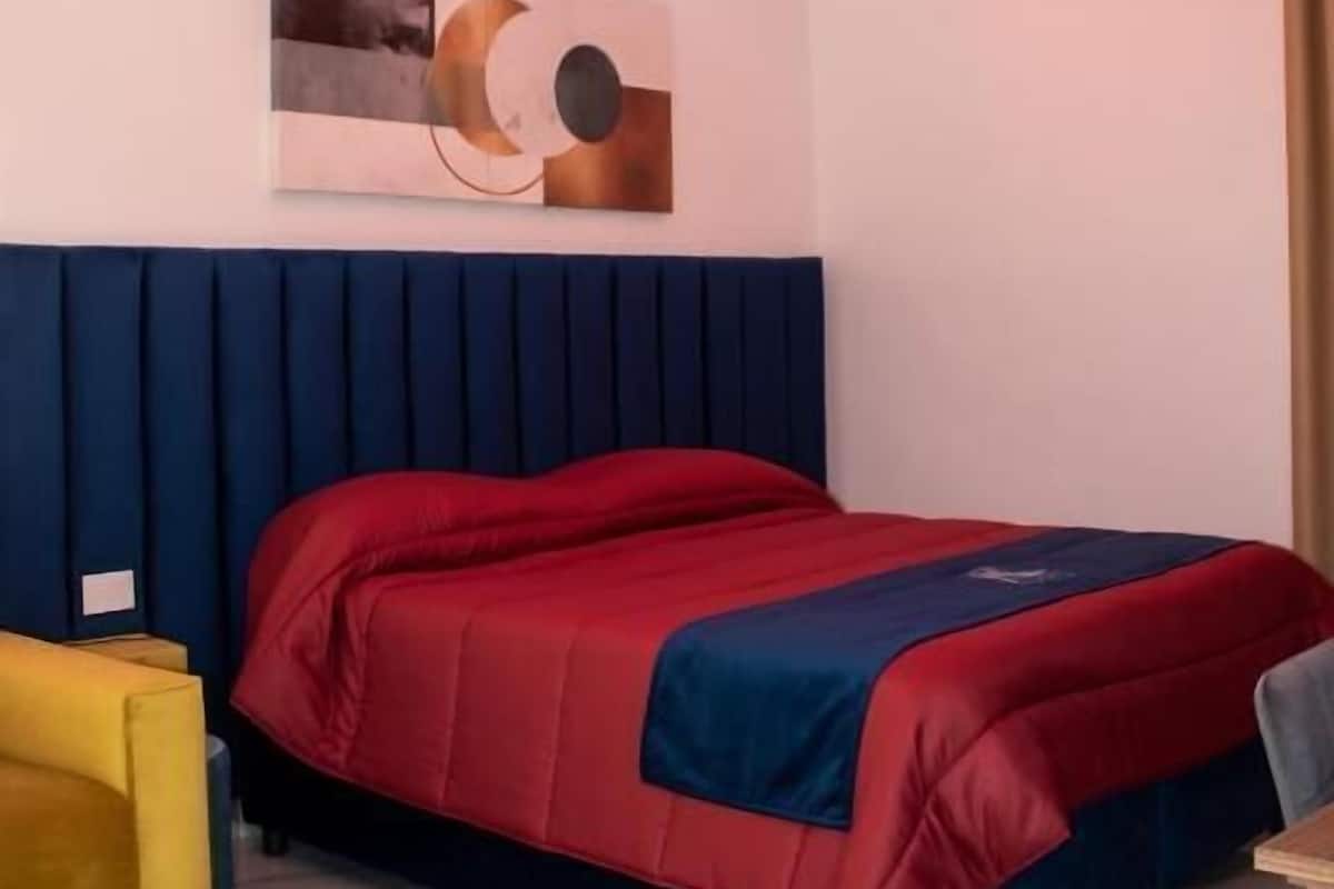 Quarto com cama, cabeceira, quadro na parede e cadeira amarela.