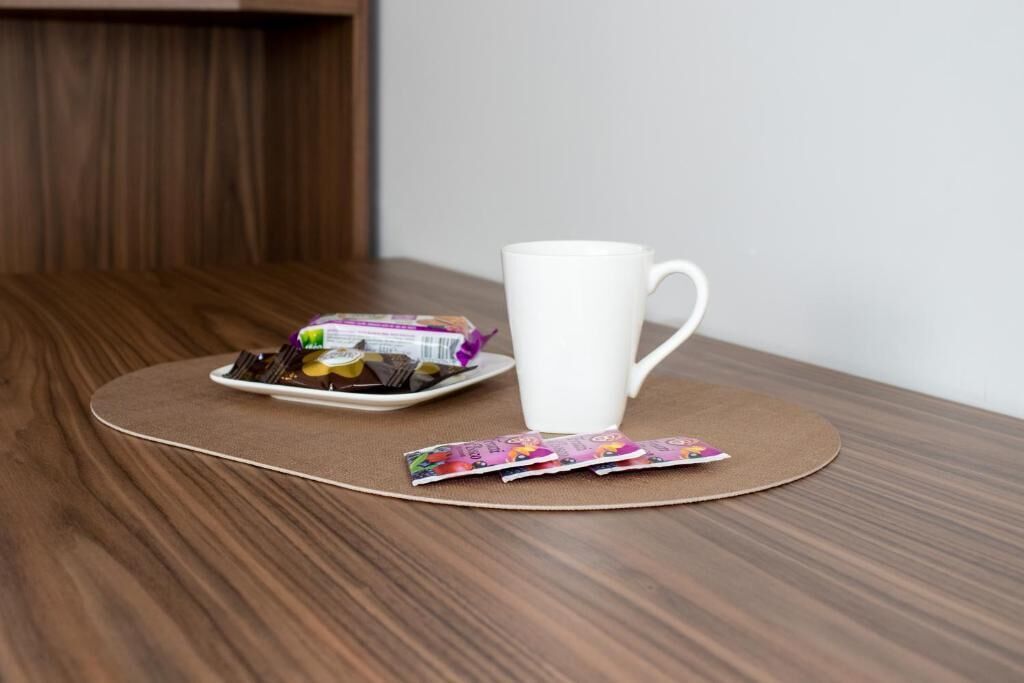 Een witte mok op een ronde tafel met een onderzetter, een bord met eten en een pakje snacks.