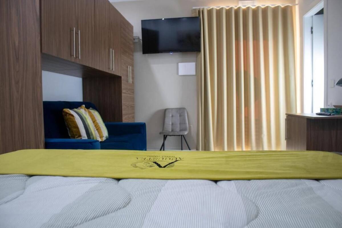 Quarto com cama, sofá azul, poltrona cinza, televisão e cortina.