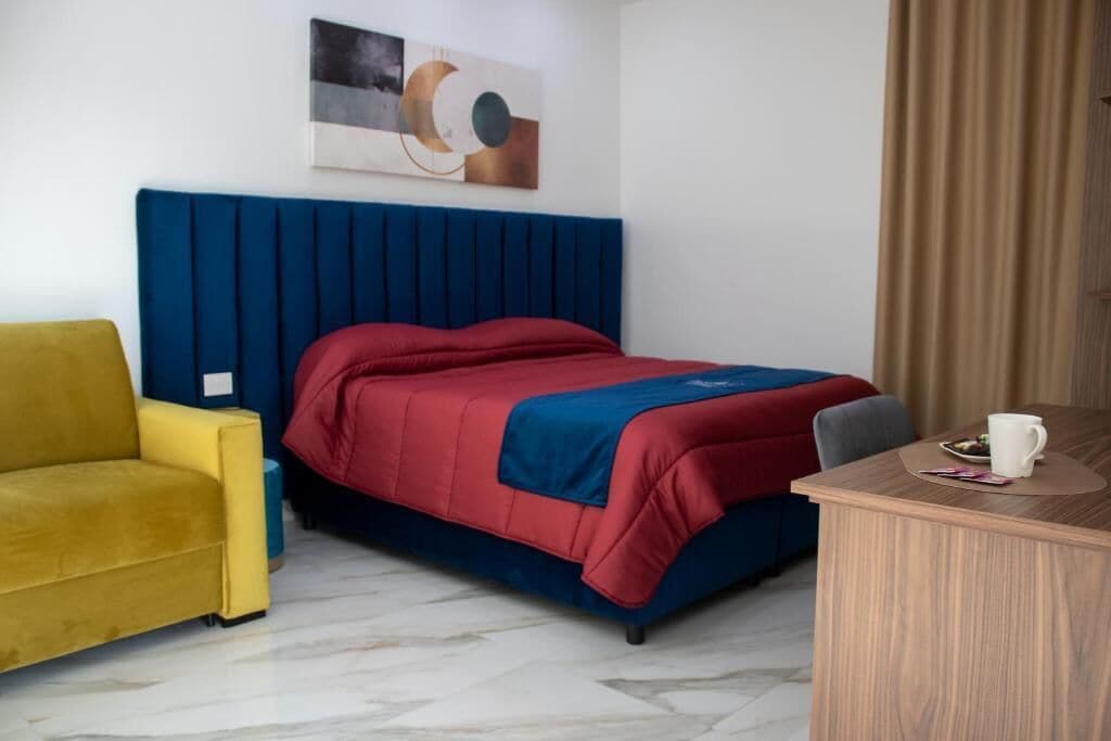 A hotel room with a bed, a yellow armchair, a wooden desk, and a painting on the wall.