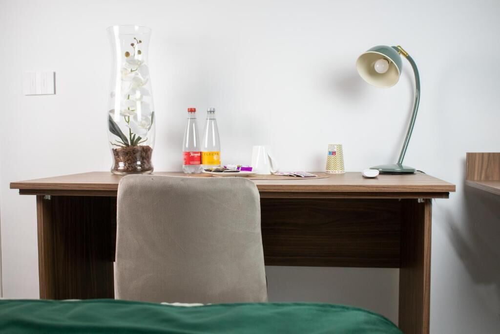 Habitación de hotel con un escritorio de madera, una silla verde, un florero con flores, dos botellas, una lámpara y una caja pequeña.