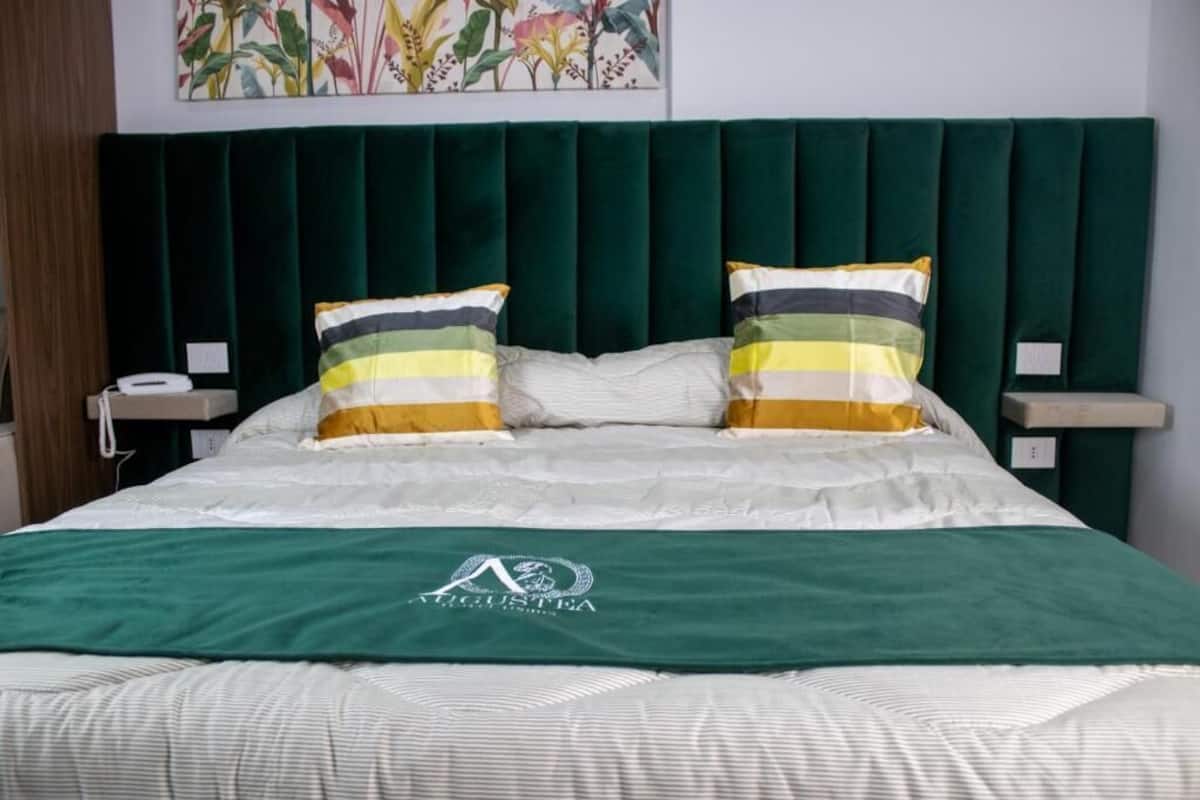 Cama bem arrumada com cabeceira verde, duas almofadas listradas e um cobertor verde com logotipo.