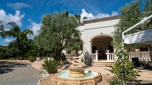 A courtyard with a fountain, palm trees, and a building with an archway. - Villa Tonia - Stanza Edera Apartment with Private Terrace, Wi-Fi, and Air Conditioning (Fasano)