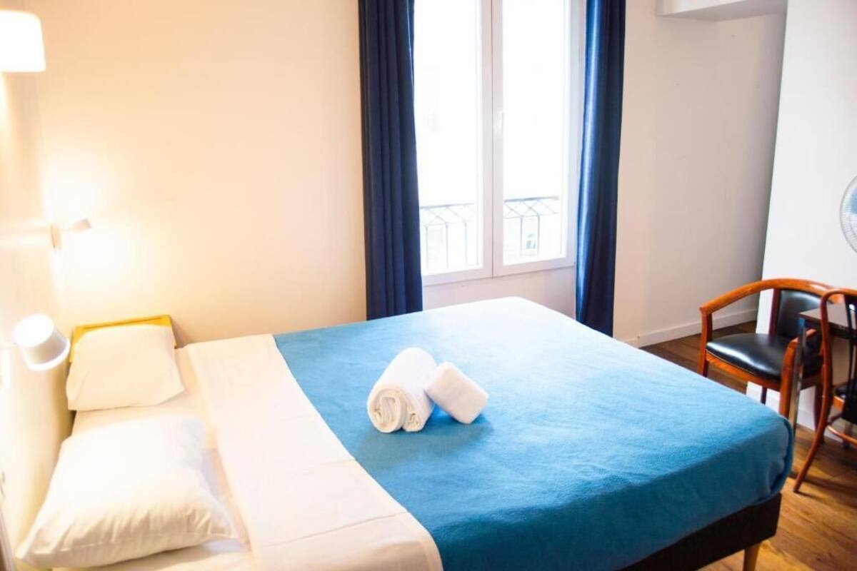 A hotel room with a bed, a chair, a window with curtains, and a wall-mounted light.