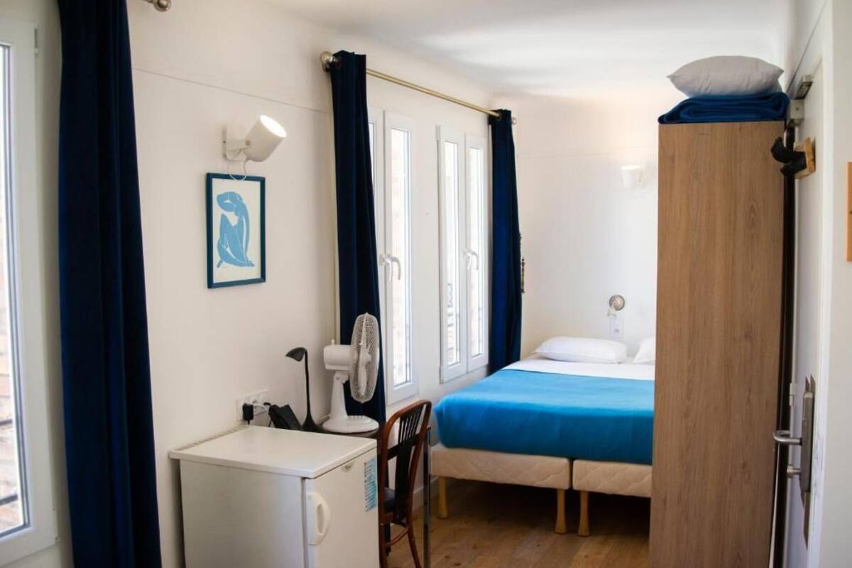 A compact room with a bed, a desk, a chair, a small fridge, and a fan.