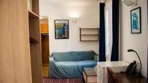 A compact room with a bed, a desk, a chair, a small table, a shelf, a poster on the wall, and a window with curtains. - HOTEL LA LOUISIANE (Paris)