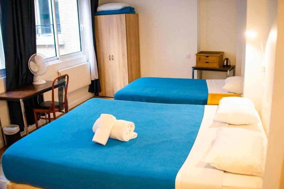 A hotel room with two beds, a desk with a chair, a fan, a wardrobe, and a small table with a box.