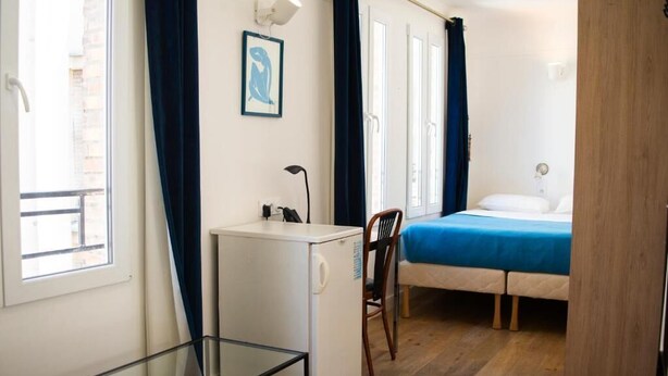 A compact room with a bed, a desk, a chair, a small refrigerator, and a wardrobe. - HOTEL LA LOUISIANE (Paris)