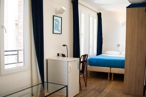 A compact room with a bed, a desk, a chair, a small refrigerator, and a wardrobe. - HOTEL LA LOUISIANE (Paris)